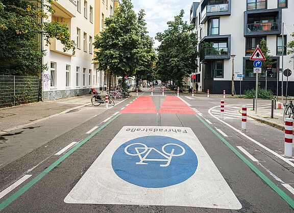 Fahrradstraße Berlin Eine Straße in Berlin: Rechts und links sind Häuserzeilen zu sehen, die Fahrbahn ist mit einem großen Fahrradsymbol versehen. Weiter hinten ist die Fahrbahn rot eingefärbt, Poller dienen als modaler Filter.